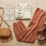 Complete boho chic outfit flat lay with cream peasant blouse, terracotta wide-leg pants, brown ankle boots and fringe bag on light wood floor