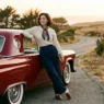 Woman in retro-inspired outfit leaning against vintage car door in warm afternoon light
