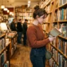 Woman in casual outfit browsing bookshelf in warm independent bookstore with soft lighting