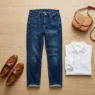 Complete casual outfit flat lay showing dark jeans, white blouse, cognac loafers and crossbody bag on wood floor