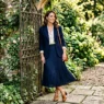 Woman in navy pleated midi skirt and cream silk blouse with structured blazer standing by garden gate