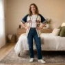 Woman in softly lit bedroom wearing styled baseball jersey outfit with confident pose and hands on hips