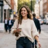 Woman wearing high-waisted jeans and oversized cream sweater with gold necklace walking on city street
