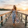 Woman in flowing summer dress walking on wooden pier over water during golden hour
