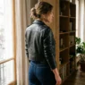 Woman in fitted black leather moto jacket standing near window with soft natural light showing full shoulder-to-hip silhouette