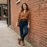 Woman in brown shirt outfit with dark denim and cognac boots leaning against a red brick wall in soft daylight