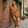 Woman in matching caramel linen wide-leg trousers and longline top with coordinating hijab leaning against red brick wall