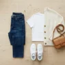 Overhead view of basic casual outfit with dark jeans, white tee, cream cardigan and white sneakers on wood floor