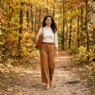 Woman in caramel wide-leg trousers and ivory mock-neck walking a soft dirt forest path in dappled autumn sunlight