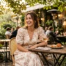 Woman in floral midi dress seated at outdoor café terrace with rosé glass and pastries in warm afternoon light