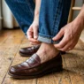 Close-up of hands precisely rolling denim hem into cuff with loafer and wood floor visible