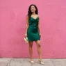 Woman in deep emerald satin cowl-neck mini dress with gold metallic mules against a vibrant pink wall