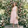 Woman in soft blush one-shoulder midi dress with ruffle hem posed in front of rose floral wall