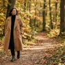 Woman in oversized camel double-breasted coat walking a forest path in autumn dappled sunlight