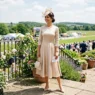 Woman in champagne A-line midi dress and cream saucer hat standing on sunny outdoor terrace