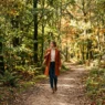 Woman walking along a dirt forest path in a styled fall outfit with dappled sunlight through autumn leaves
