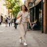 Woman walking on a sunlit city sidewalk in an oatmeal linen co-ord set with wide-leg trousers and white sneakers