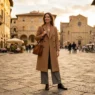 Woman in camel double-breasted wool overcoat and wide-leg grey trousers on European cobblestone piazza in golden afternoon light