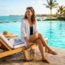 Woman in chic swimsuit coverup and sandals seated on a sunlit pool deck lounger with turquoise water behind her