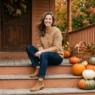 Woman in camel knit sweater and ankle boots seated on painted porch steps with pumpkins in soft autumn light