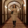 Woman in beige trench coat belted over slim black trousers and black loafers in hotel corridor with warm sconce lighting