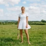 Woman standing in a grass field wearing a crisp white tennis dress with a flared skirt and white sneakers in bright daylight